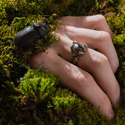 A hand wearing the Rhinoceros Beetle Ring, resting on moss with a real beetle nearby. The organic setting highlights the nature-inspired design of the jewelry, blending seamlessly with its surroundings.