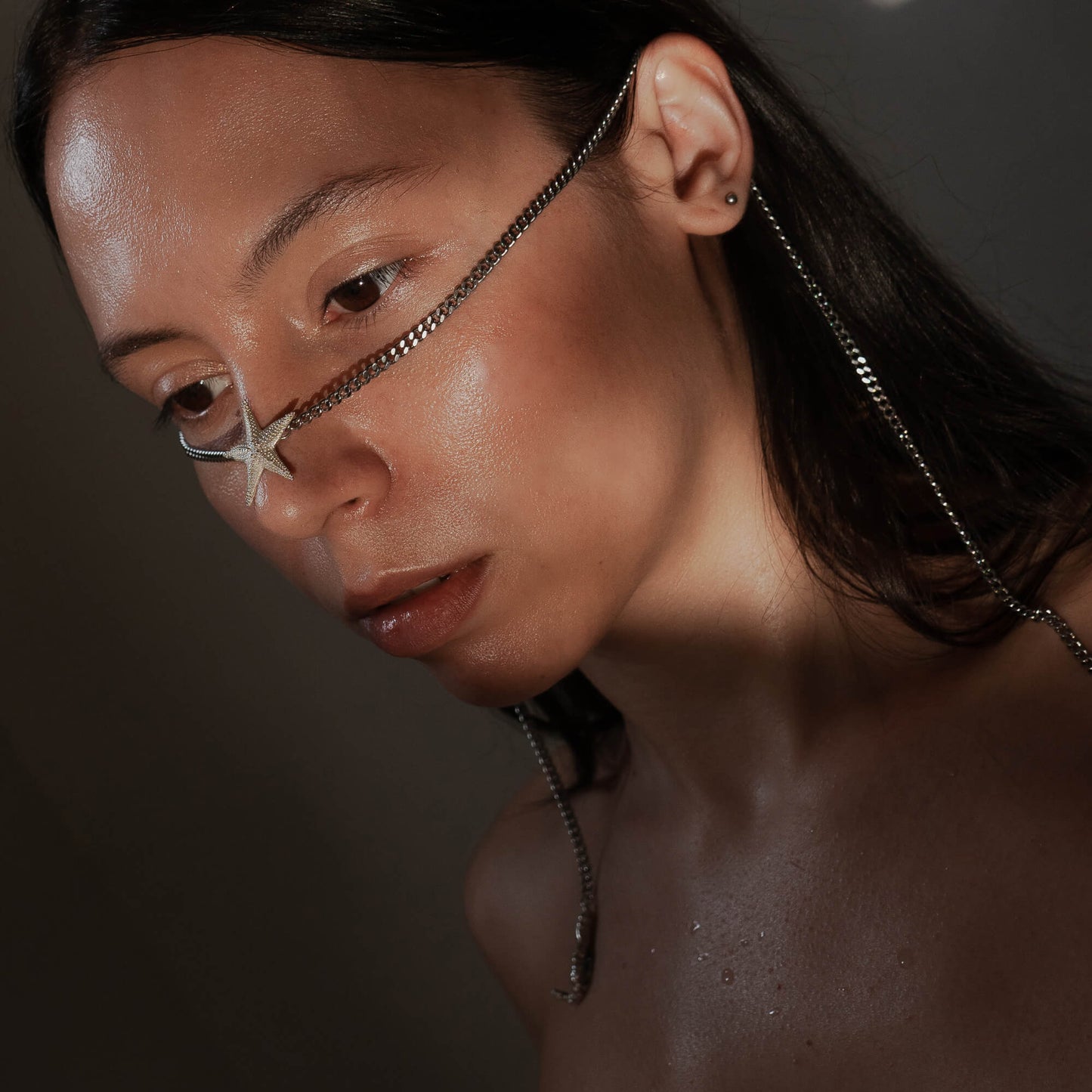 A close-up of a model with wet, glowing skin wearing the Starfish Facechain. The silver chain wraps around the face, with a starfish detail on the nose and pearl-adorned ends dangling at the sides.