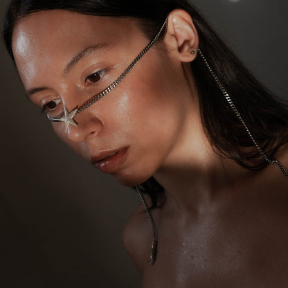 A close-up of a model with wet, glowing skin wearing the Starfish Facechain. The silver chain wraps around the face, with a starfish detail on the nose and pearl-adorned ends dangling at the sides.